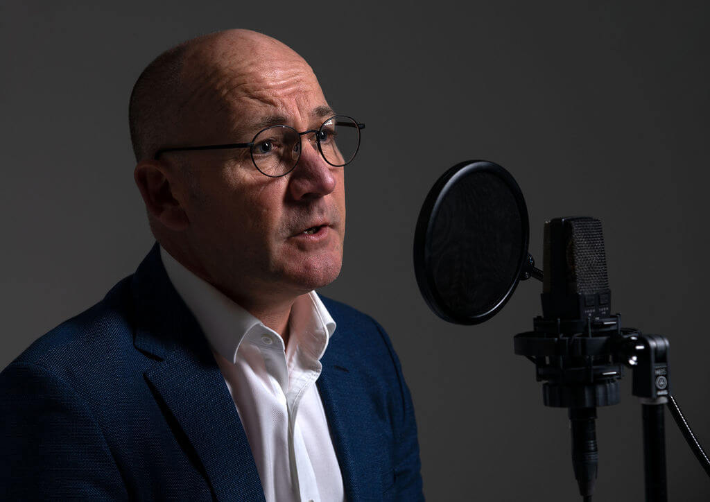 James Fowler voiceover talking into a microphone whilst looking thoughtful and wearing a suit.