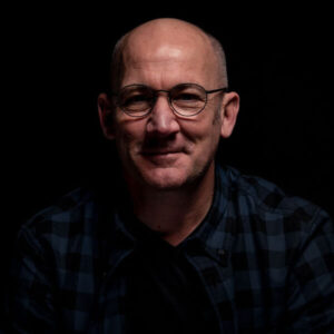 James Fowler voiceover headshot with very dark background. James is smiling, looking into the camera.