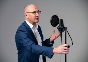 James Fowler Voiceover talking into a microphone wearing a blue suit and white shirt.